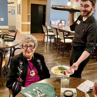 A senior woman sits at a table in a restaurant while a waiter serves her meal.