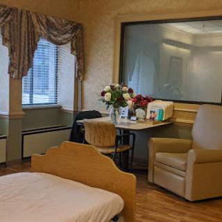 A peaceful hospital room featuring a cozy chair, a table with flowers, and sunlight streaming in through the window.