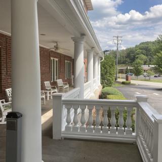 A sunny view of a porch with columns, chairs, and a brick building.