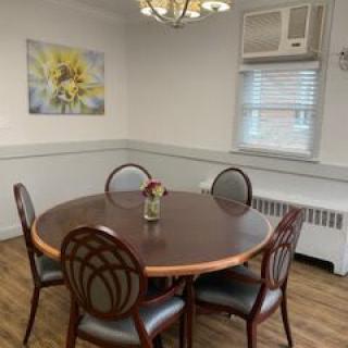 A small dining room with a round table and four chairs, featuring a wall painting and window with an air conditioning unit.