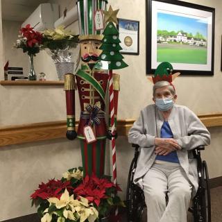 An elderly person in a wheelchair sits beside a festive nutcracker and poinsettias, wearing a mask in an indoor setting decorated for Christmas.
