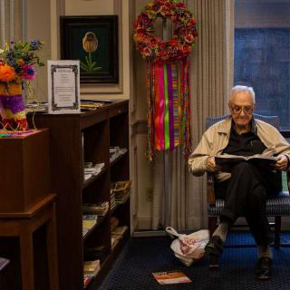 An elderly person sitting in a room, reading a newspaper next to a decorated bookshelf.