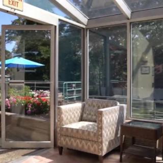 A cozy lounge area with a patterned chair, surrounded by large windows showcasing a sunny outdoor patio.