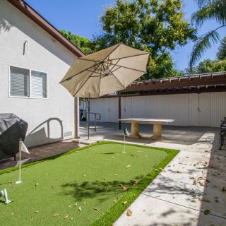 A sunny backyard with a small putting green, table tennis setup under an umbrella, and a relaxing patio area.
