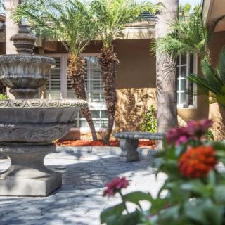 A tranquil courtyard featuring a stone fountain surrounded by palm trees and colorful flowers.