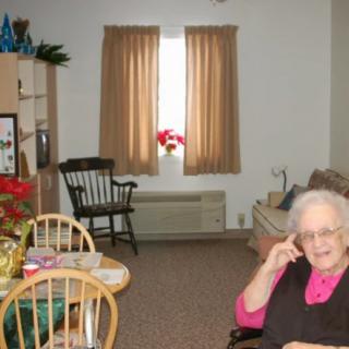 A senior woman sits in a cozy living room with simple furnishings.
