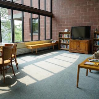 A bright and inviting living room with large windows allowing natural light to fill the space, featuring a collection of furniture including a bookshelf and a television.