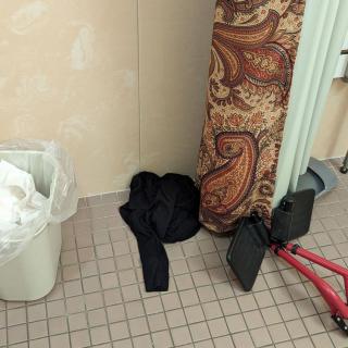 A cluttered corner of a bathroom with a coat, trash can, carpet, and exercise equipment.