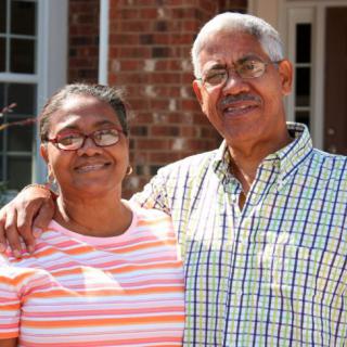 An elderly couple standing outside in front of their home.