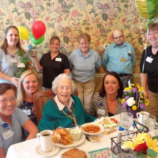 A joyful celebration with family and friends, featuring a senior woman at the center of attention, surrounded by balloons and flowers.