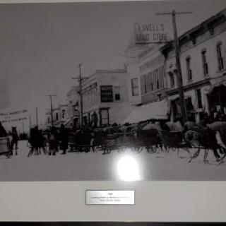 A historical black and white photograph depicting a busy street scene with horse-drawn carriages and pedestrians in front of early 20th-century buildings.
