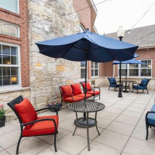 A cozy outdoor patio area featuring comfortable chairs with red cushions, blue umbrellas, and metal tables against a brick building.