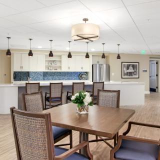 A modern dining area with a wooden table and upholstered chairs, adjacent to a spacious kitchen featuring pendant lights.