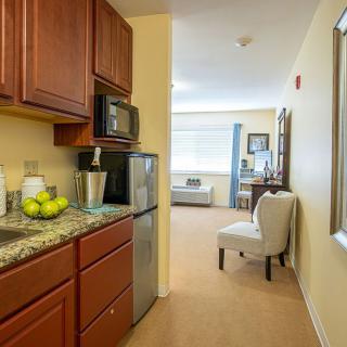 A cozy apartment interior featuring a compact kitchen area with a fridge and microwave, adjacent to a small living space with a chair.