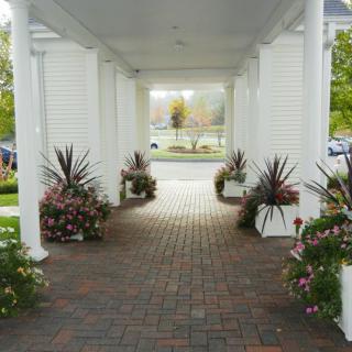 A covered walkway with white columns and brick flooring, adorned with potted plants and flowers.