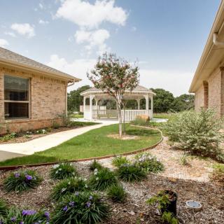 A scenic garden view featuring a white gazebo, bordered by brick buildings and blooming flowers under a sunny sky.