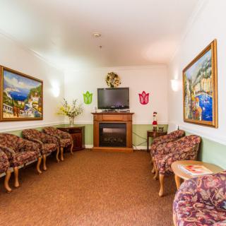 A cozy living room with a television above a fireplace, surrounded by comfortable seating and artwork on the walls.