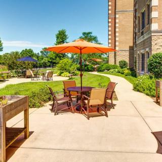 A sunny outdoor patio area with tables and chairs under an orange umbrella, surrounded by greenery and a building.