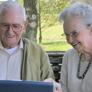 An elderly couple smiling while looking at a laptop outdoors.