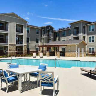 A modern apartment complex featuring a swimming pool and outdoor seating area under a clear blue sky.