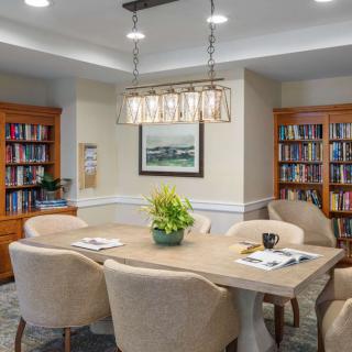 A cozy library room with bookshelves, a large table surrounded by chairs, and a modern light fixture above.