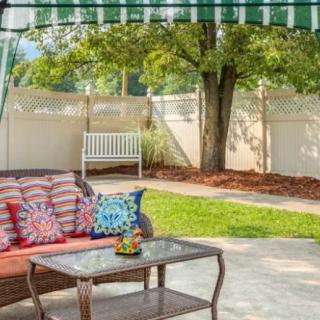A cozy outdoor patio with a sofa, colorful pillows, and a table underneath an awning, surrounded by a garden with trees and a fence.