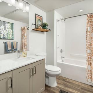 A modern bathroom featuring a vanity with a large mirror, sink, and storage cabinets. It includes a shower with a patterned curtain and overhead lighting.