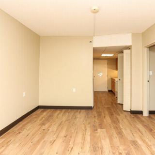 An empty apartment room with wood flooring and a view into a hallway.