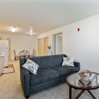 A cozy apartment living room with a sofa, table, and an open view of the kitchen and dining area.