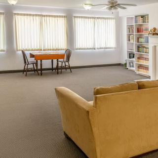 A spacious living room with a beige sofa, bookshelf, and a small dining table near the windows.