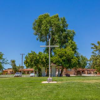 A cross stands on a grassy area with a building and trees in the background.