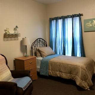 A cozy bedroom with a single bed, side table, and a comfortable chair next to a window with blue curtains.