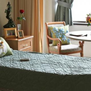 A cozy bedroom with a neatly arranged bed, a wooden chair by the window, and a small table adorned with flowers.