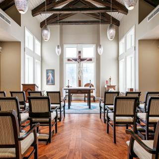 A serene chapel interior featuring rows of chairs facing an altar with a prominent cross and large windows letting in natural light.