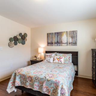 A cozy bedroom with a floral bedspread, artwork on the wall, and a warm lamp on the bedside table.