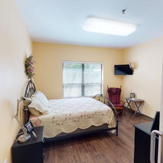 A cozy bedroom featuring a bed, nightstand, wall-mounted television, and a chair by the window.