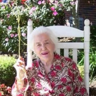 An elderly woman sitting in a rocking chair in a sunny garden, surrounded by flowers.