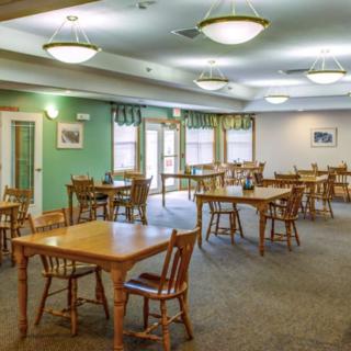 A spacious dining room with multiple tables and chairs, well-lit with ceiling lights and natural light from the windows.