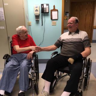 Two elderly men in wheelchairs shaking hands warmly in a nursing home.