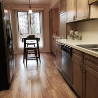 A modern kitchen with wooden flooring, featuring appliances and a small dining area by the window.
