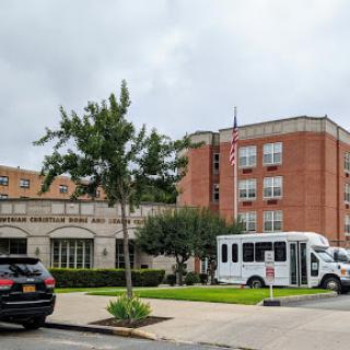 A view of a hospital building with parked vehicles and a transportation shuttle in the foreground.