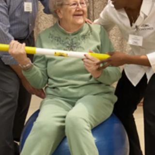 An elderly person engaging in physical therapy with assistance, sitting on a stability ball and holding a rod.