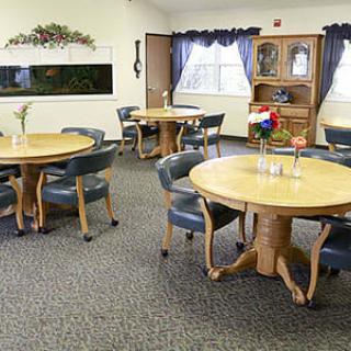 A cozy dining area with several round tables and chairs arranged on a carpeted floor, featuring windows with curtains and decorative accents.