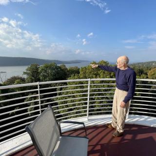 A man stands on a balcony overlooking a scenic view of a lake and trees.