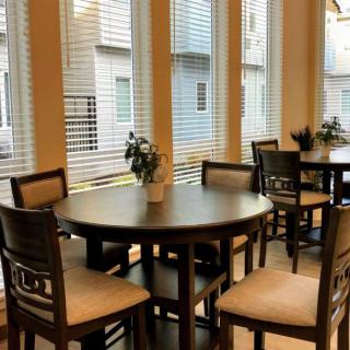 A cozy dining area with round tables and chairs, illuminated by natural light through large windows with blinds.