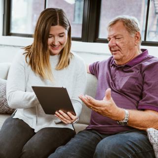 A young woman shows an older man something on a tablet while sitting on a couch.