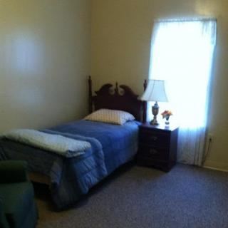 A cozy, simple bedroom featuring a single bed, a nightstand with a lamp, and natural light coming through the window.