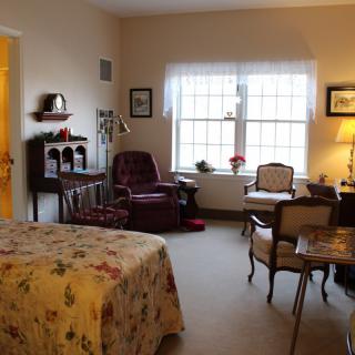 A cozy bedroom interior with traditional furnishings, featuring a bed, armchair, and a desk near a large window.