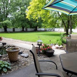 A peaceful backyard garden with a patio area, featuring chairs, an umbrella, and potted plants under lush green trees.