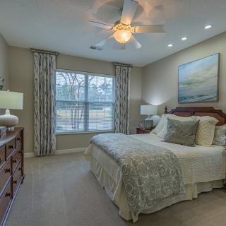 A cozy bedroom with a well-made bed, nightstands, and lamps, featuring a large window with curtains and a ceiling fan overhead.